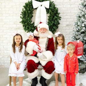 May include: Santa Claus sits on a bench with four children. The children are wearing white pajamas with red trim. Santa is wearing a traditional red suit with white fur trim. There is a wreath with a white bow behind Santa and a red mailbox with "North Pole Post" written on it to the right.