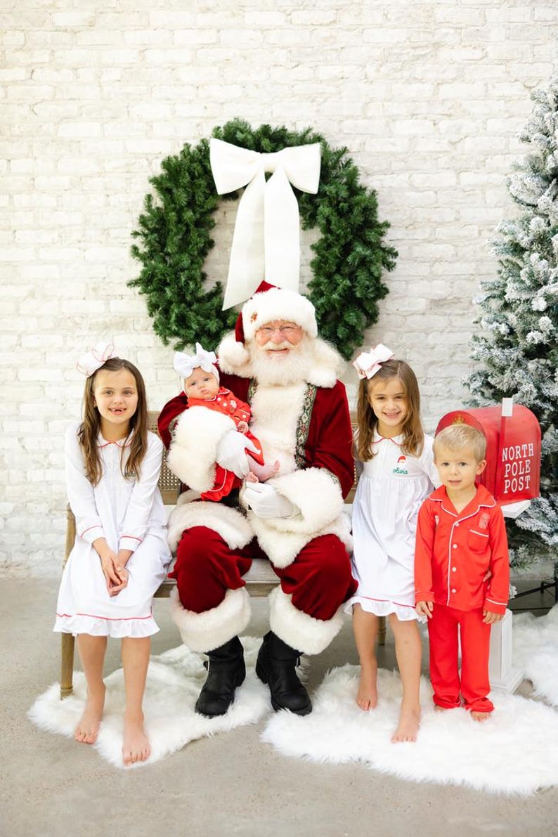 May include: Santa Claus sits on a bench with four children. The children are wearing white pajamas with red trim. Santa is wearing a traditional red suit with white fur trim. There is a wreath with a white bow behind Santa and a red mailbox with "North Pole Post" written on it to the right.