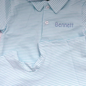 May include: A short-sleeved polo shirt with a light blue and white horizontal striped pattern. The shirt features a collar, two white buttons, and the name Bennett embroidered in blue thread on the chest. A tag labelled Mila & Olivia Designs is visible.