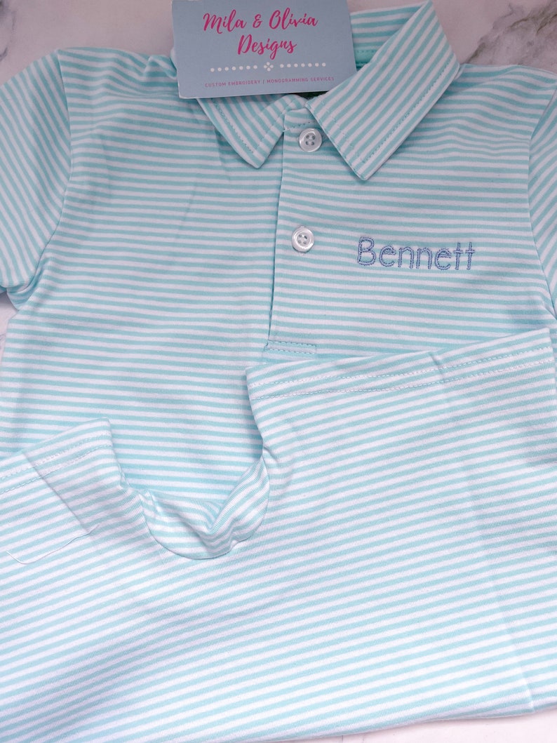 May include: A short-sleeved polo shirt with a light blue and white horizontal striped pattern. The shirt features a collar, two white buttons, and the name Bennett embroidered in blue thread on the chest. A tag labelled Mila & Olivia Designs is visible.