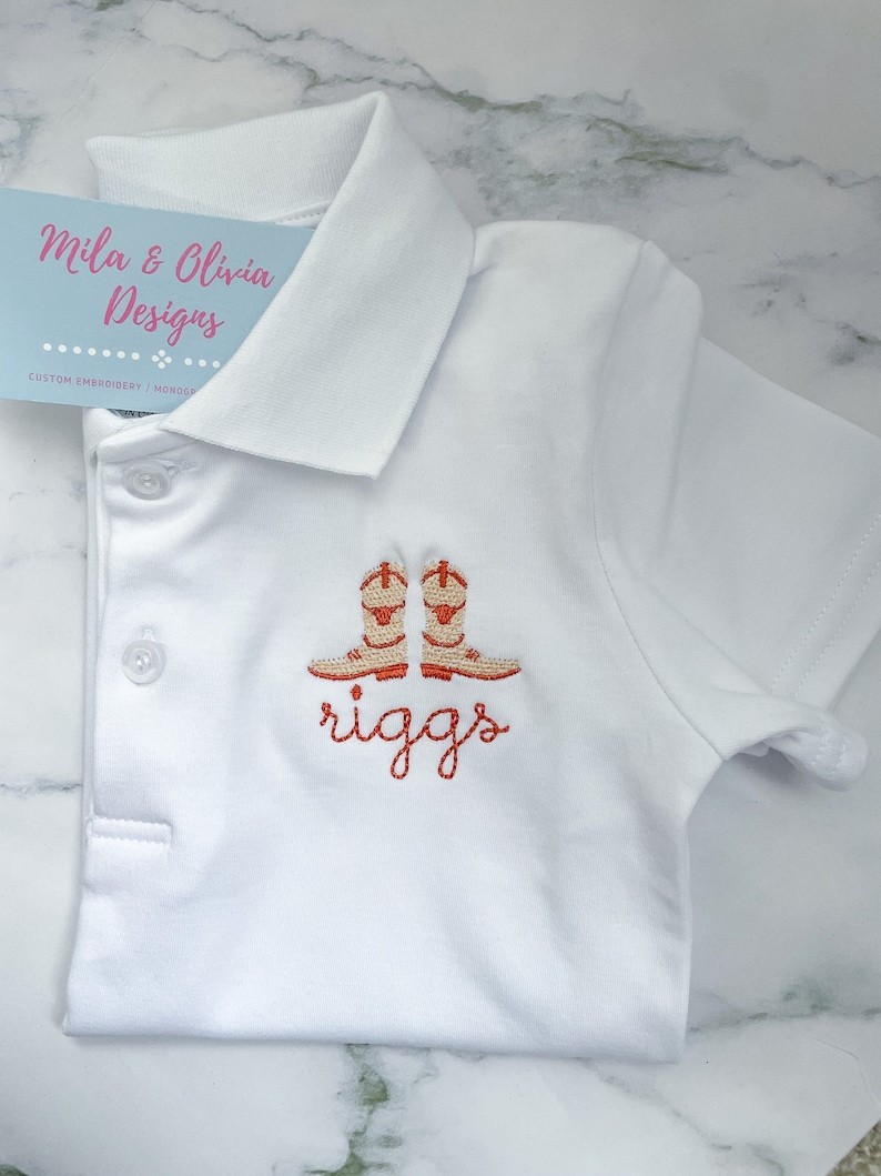 K&ouml;nnte beinhalten: Wei&szlig;es Poloshirt mit orangefarbenen gestickten Cowboystiefeln und dem Namen "riggs" darunter.