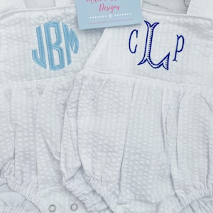 May include: Two white baby onesies with embroidered monograms. The onesie on the left has a light blue monogram "JBM" and the onesie on the right has a dark blue monogram "CLP". The onesies are on a white marble surface. The text "Mila & Olivia Designs" is on a white card with pink lettering.