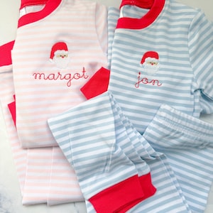 May include: Two sets of children's pajamas. One set is light pink with white stripes, the other is light blue with white stripes. Both sets have red trim and a Santa Claus embroidery. The pink set says "margot" and the blue set says "jon".