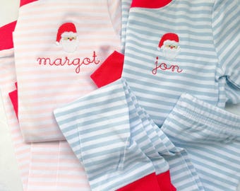 May include: Two sets of children's pajamas. One set is light pink with white stripes, the other is light blue with white stripes. Both sets have red trim and a Santa Claus embroidery. The pink set says "margot" and the blue set says "jon".