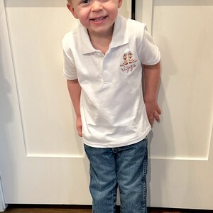 K&ouml;nnte beinhalten: Ein kleiner Junge tr&auml;gt ein wei&szlig;es Poloshirt mit einem braunen gestickten Design von Cowboystiefeln und dem Wort "riggs". Er tr&auml;gt eine blaue Jeans und braune Cowboystiefel. Er steht vor einer wei&szlig;en T&uuml;r.