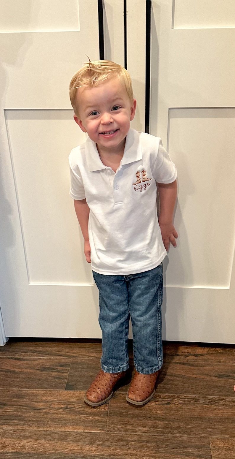 K&ouml;nnte beinhalten: Ein kleiner Junge tr&auml;gt ein wei&szlig;es Poloshirt mit einem braunen gestickten Design von Cowboystiefeln und dem Wort "riggs". Er tr&auml;gt eine blaue Jeans und braune Cowboystiefel. Er steht vor einer wei&szlig;en T&uuml;r.