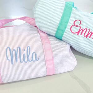 May include: Two duffel bags made of seersucker fabric. One bag is white and light pink with pink straps and the name Mila embroidered in light blue. The other bag is white and light blue with teal straps and the name Emma embroidered in hot pink.