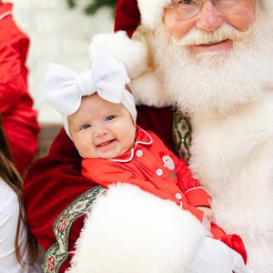 May include: Santa Claus wearing a red suit and white beard holds a baby wearing a red and white onesie and a white bow.