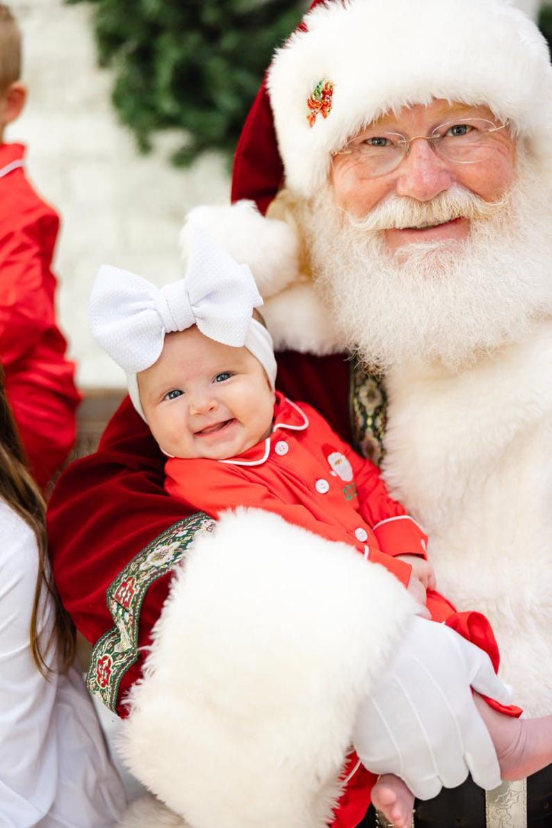 May include: Santa Claus wearing a red suit and white beard holds a baby wearing a red and white onesie and a white bow.