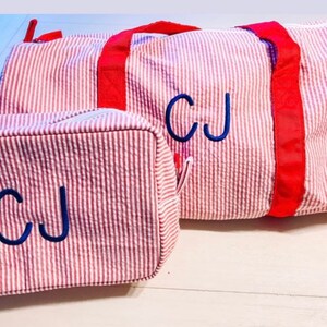 May include: A red and white seersucker duffel bag and a matching rectangular toiletry bag. Both items feature bright red fabric straps and zippers, with the initials CJ embroidered in dark blue thread on the front of each piece.