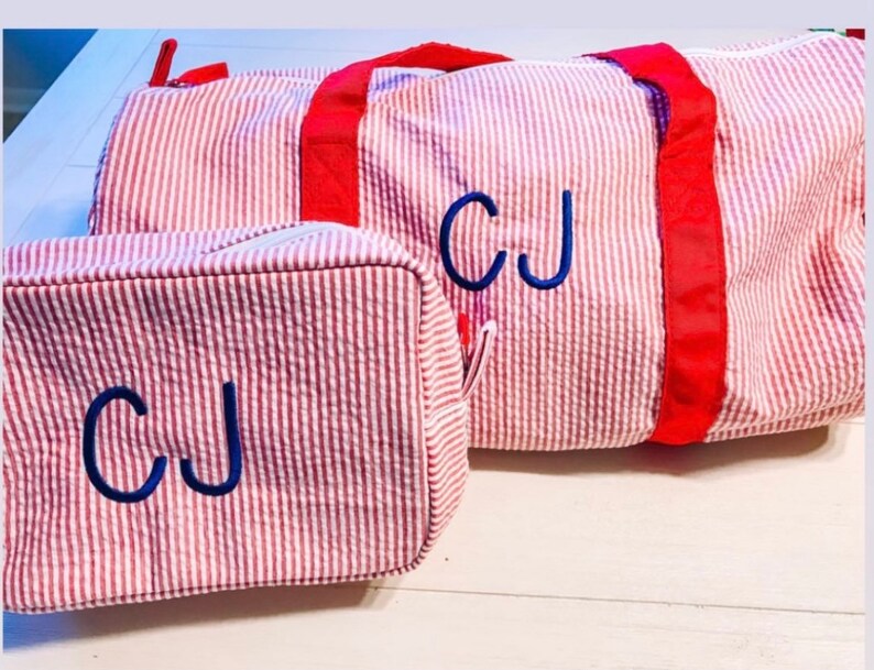 May include: A red and white seersucker duffel bag and a matching rectangular toiletry bag. Both items feature bright red fabric straps and zippers, with the initials CJ embroidered in dark blue thread on the front of each piece.