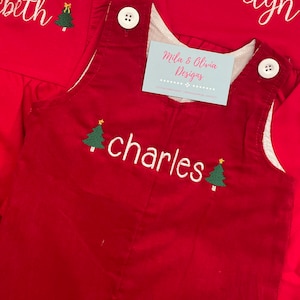 May include: Red corduroy overalls with white straps and white stitching. The overalls have a white embroidered name "charles" and two green embroidered Christmas trees with yellow stars. A small blue and white business card with the text "Mila & Olivia Designs" is visible in the image.