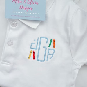 May include: White polo shirt with a blue embroidered letter G and colorful crayons around it. The shirt has a small label that says "Mila & Olivia Designs, Custom Embroidery / Monogramming Services".