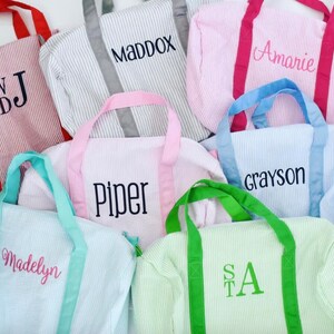 May include: A collection of seersucker duffel bags in various colors including red, gray, pink, light blue, and green. Each bag features contrasting handles and is personalized with embroidered names or initials such as WDJ, MADDOX, Amarie, Piper, Grayson, Madelyn, and STA.