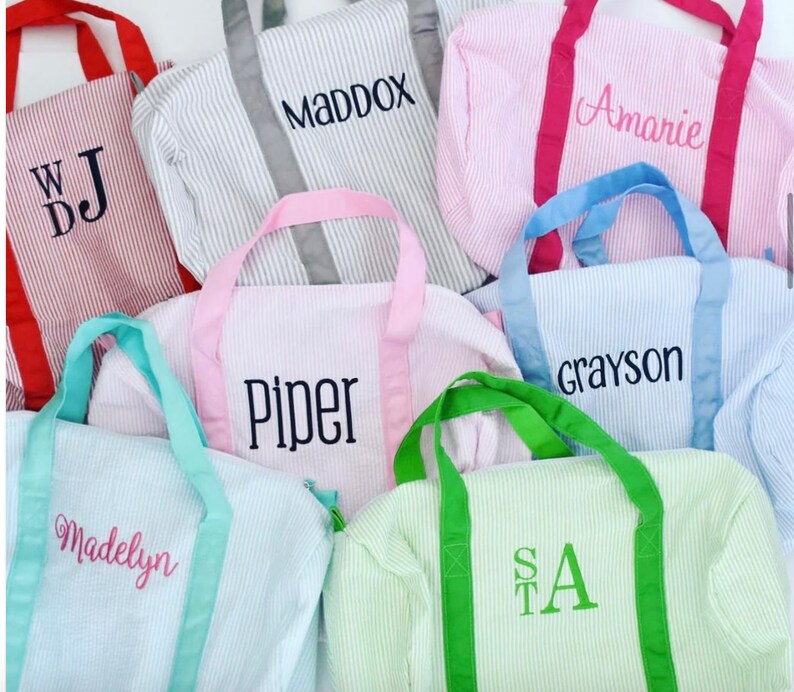 May include: A collection of seersucker duffel bags in various colors including red, gray, pink, light blue, and green. Each bag features contrasting handles and is personalized with embroidered names or initials such as WDJ, MADDOX, Amarie, Piper, Grayson, Madelyn, and STA.