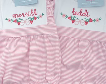 May include: Two pink and white striped baby onesies with embroidered strawberry designs and personalized names "merritt" and "teddi".