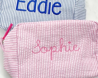 May include: Two rectangular zippered pouches made of seersucker fabric. One pouch features blue and white vertical stripes with the name Eddie embroidered in blue. The other pouch features pink and white vertical stripes with the name Sophie embroidered in pink.