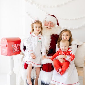 May include: Santa Claus is sitting on a white couch with three children. The children are wearing red and white pajamas. A red mailbox with the words "North Pole Post" is in the background.