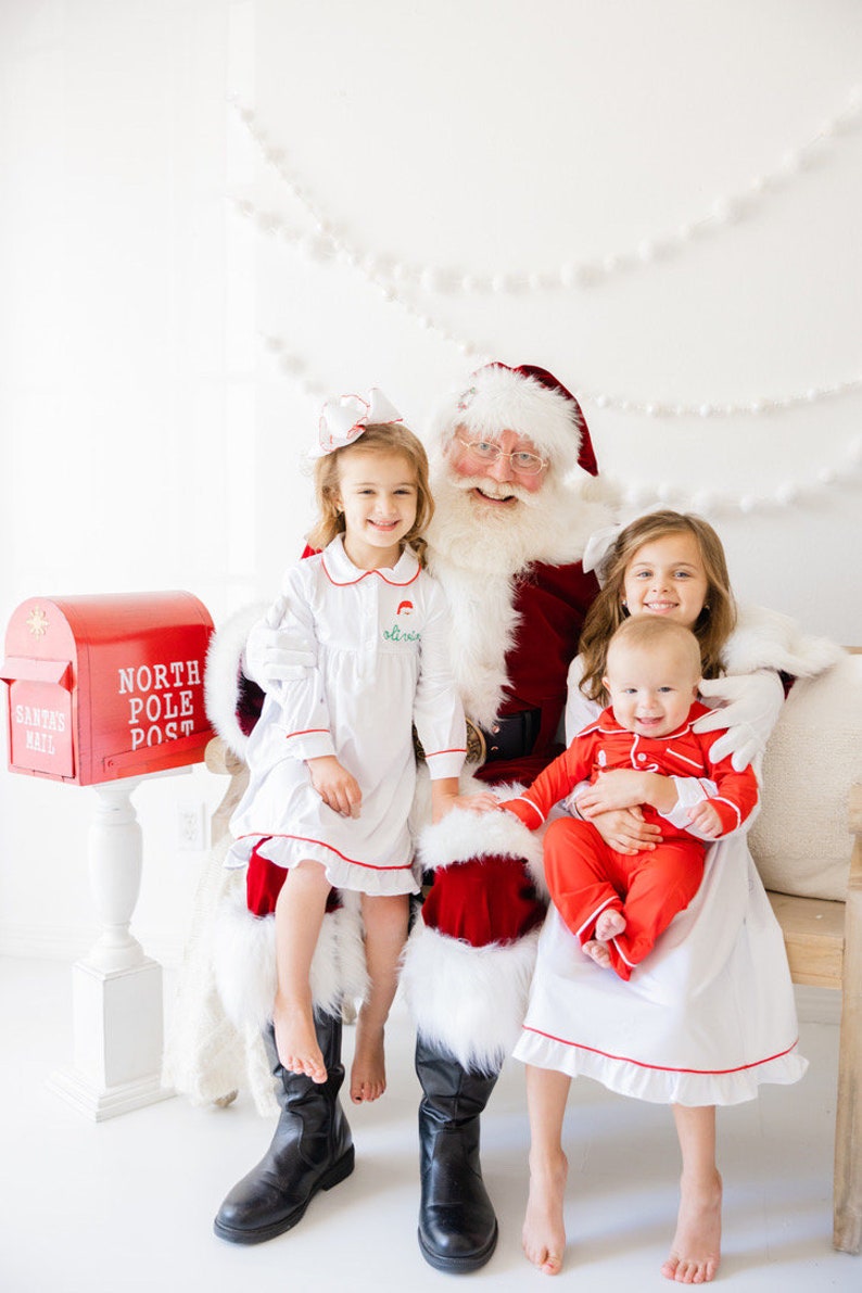 May include: Santa Claus is sitting on a white couch with three children. The children are wearing red and white pajamas. A red mailbox with the words "North Pole Post" is in the background.