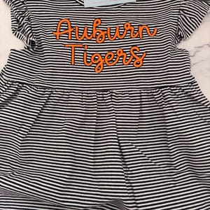 May include: A black and white striped dress with orange embroidered text that reads "Auburn Tigers." The dress has ruffled sleeves and a gathered skirt. A small card with the text "Mila & Olivia Designs" is attached.