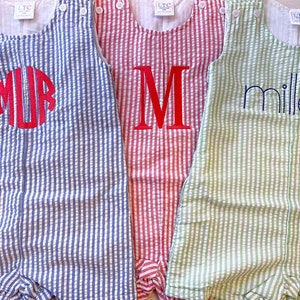 May include: Three seersucker rompers in blue, red, and green. The blue romper has a red monogram, the red romper has a red letter "M", and the green romper has the name "miller" embroidered in navy blue. Each has white buttons.