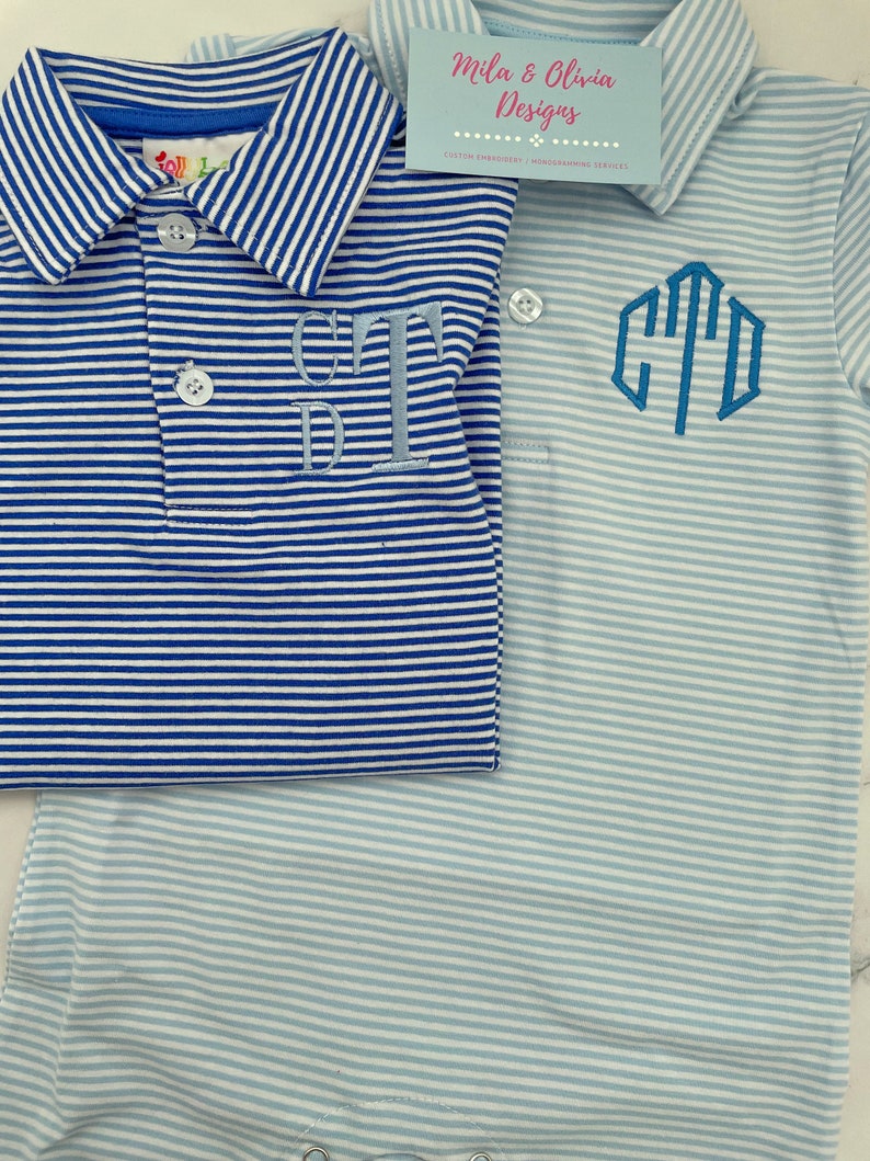 May include: Two short-sleeved polo shirts with horizontal stripes. One shirt is dark blue and white with a light blue embroidered monogram. The other is light blue and white with a dark blue embroidered monogram. A business card reads Mila & Olivia Designs.