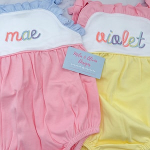 May include: Two baby rompers, one pink and one yellow, with white bibs and colorful embroidered names. The pink romper has a blue ruffle, and the yellow romper has a pink ruffle. A small card reads "Mila & Olivia Designs."