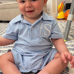 May include: A light blue and white horizontal striped short-sleeved romper featuring a collared neckline and a button placket. The garment has white embroidered monogram letters JS on the left chest area and includes metal snaps at the bottom.