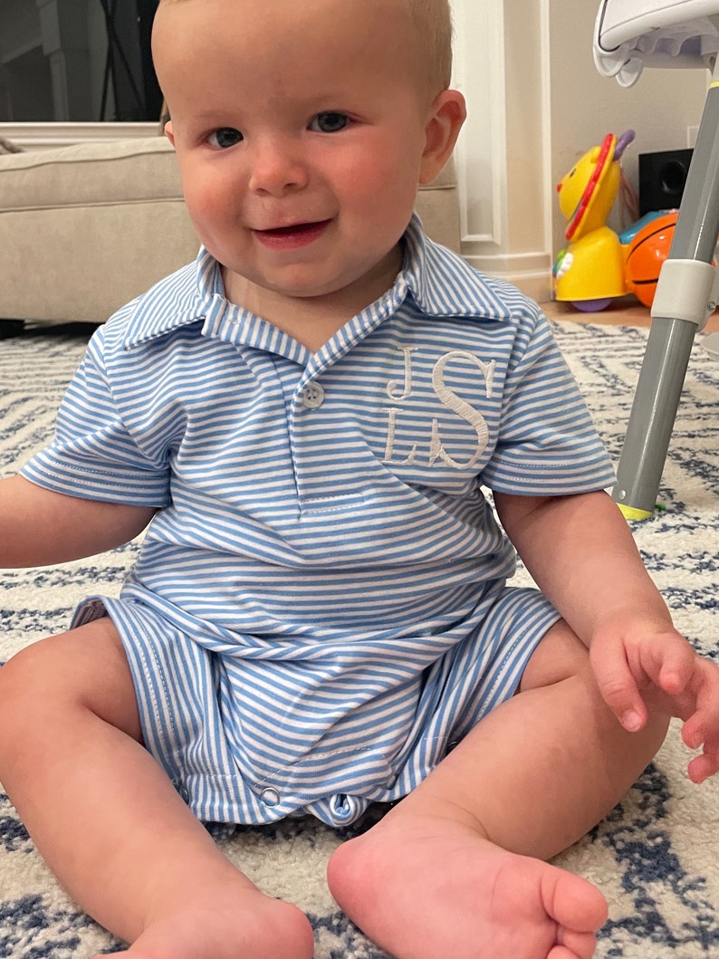 May include: A light blue and white horizontal striped short-sleeved romper featuring a collared neckline and a button placket. The garment has white embroidered monogram letters JS on the left chest area and includes metal snaps at the bottom.