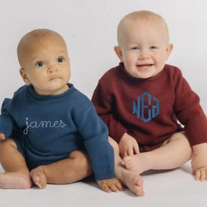 May include: Two long-sleeved knit sweaters for infants. One sweater is dark blue with the name james embroidered in white cursive script. The other sweater is maroon with a blue embroidered monogram featuring the letters NEA in a hexagonal shape.