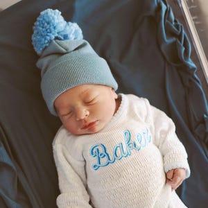 May include: A newborn baby wearing a white knitted onesie with the name "Baker" embroidered in blue. The baby is wearing a light blue beanie with a blue pom-pom on top.