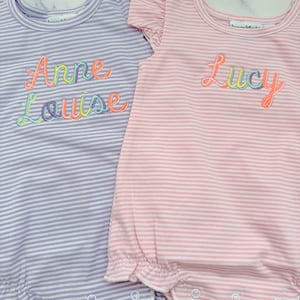 May include: Two baby rompers, one lavender and one pink, with horizontal stripes. The lavender romper has the embroidered name "Anne Louise" in rainbow colors. The pink romper has the embroidered name "Lucy" in rainbow colors.