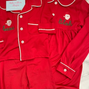 May include: Red Christmas pajamas with white trim and Santa Claus embroidery. The pajamas are personalized with the names "Beau" and "Lilah".