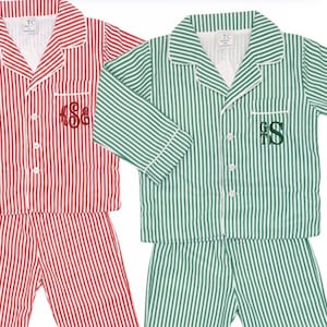 May include: Two sets of long-sleeved button-down pajamas with white piping. One set features red and white vertical stripes with a red embroidered monogram on the chest pocket. The other set has green and white vertical stripes with a green monogram.