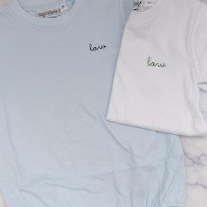 May include: Two baby rompers, one light blue and one white, are displayed. The light blue romper has the word "lane" embroidered in black, while the white romper has the same word embroidered in green. Both have the size "2T" on the tag.