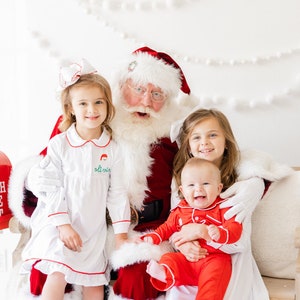 May include: Santa Claus sits with three children, two girls and a baby boy, all wearing festive pajamas. The girls are smiling and the baby is laughing. Santa is wearing a red suit with white trim and a white beard. There is a red mailbox with "North Pole Post" written on it in the background.