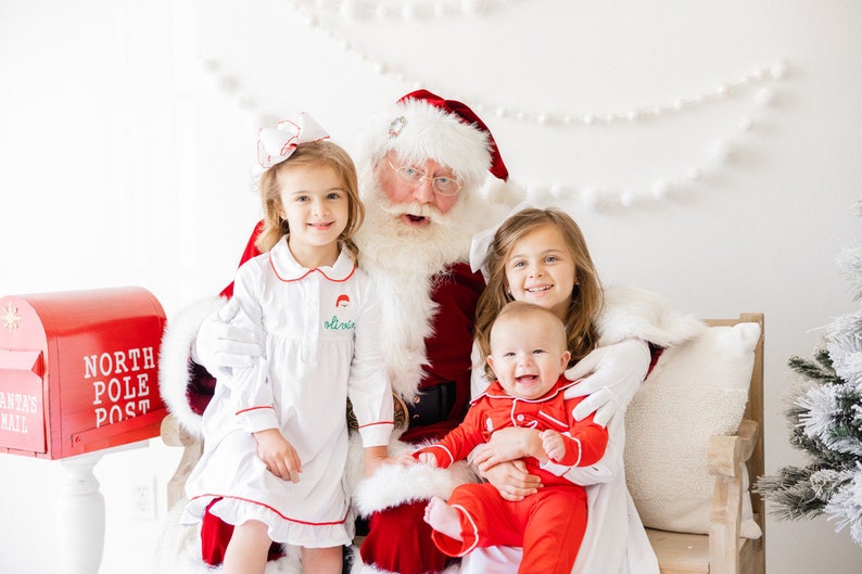 May include: Santa Claus sits with three children, two girls and a baby boy, all wearing festive pajamas. The girls are smiling and the baby is laughing. Santa is wearing a red suit with white trim and a white beard. There is a red mailbox with "North Pole Post" written on it in the background.