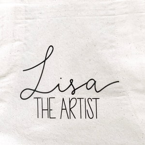 May include: White canvas tote bag with black text that reads "Lisa the Artist" in a cursive font.