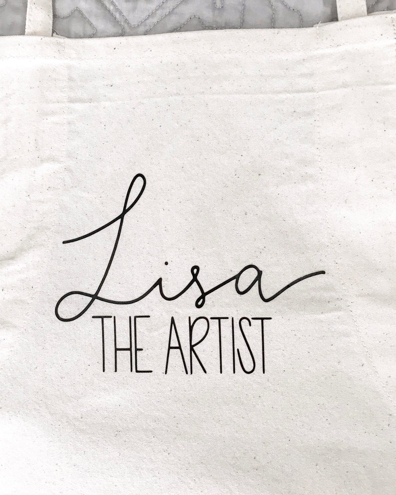 May include: White canvas tote bag with black text that reads "Lisa the Artist" in a cursive font.