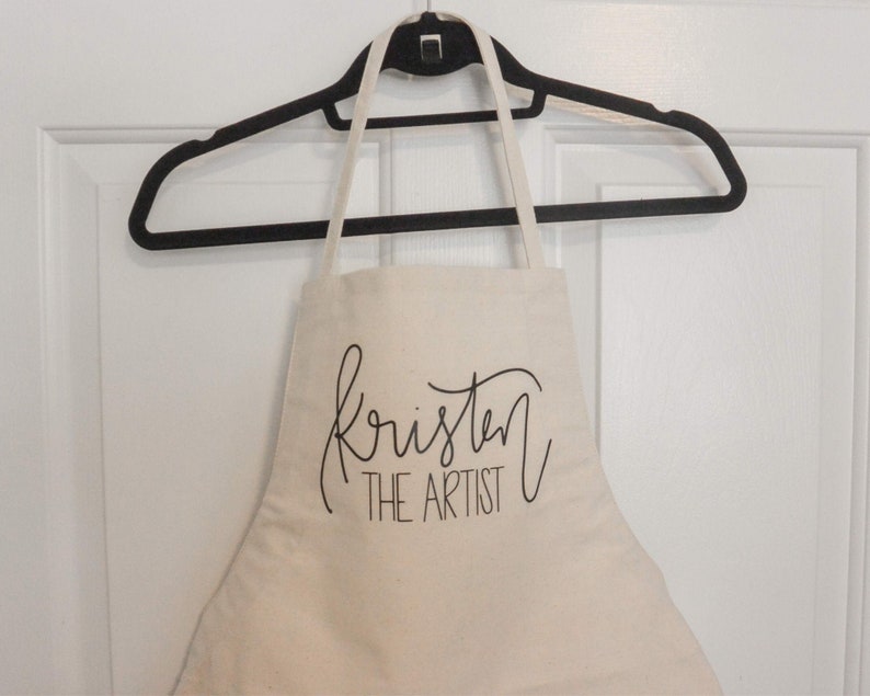 May include: A white canvas apron with black text that reads "Kristen The Artist". The apron is hanging on a black hanger.