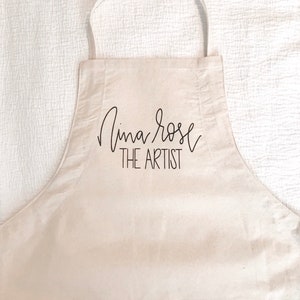 May include: A white canvas apron with a black pocket and the text "Nina Rose THE ARTIST" printed on the front.