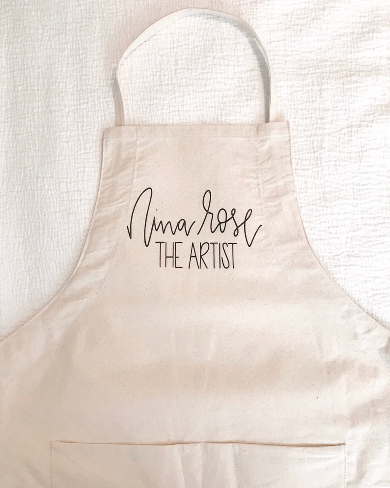 May include: A white canvas apron with a black pocket and the text "Nina Rose THE ARTIST" printed on the front.