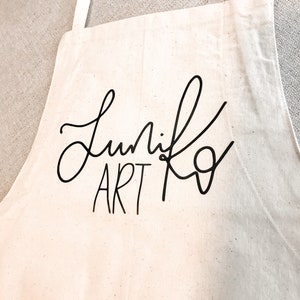 May include: A white canvas apron with black text that reads "Lunik Art" in a cursive font.