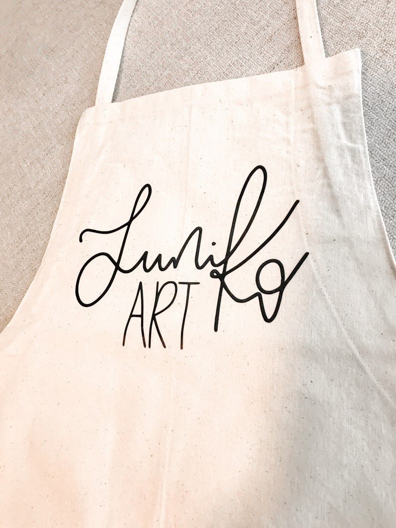 May include: A white canvas apron with black text that reads "Lunik Art" in a cursive font.