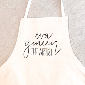 May include: White canvas apron with black text that reads "eva gineen THE ARTIST".