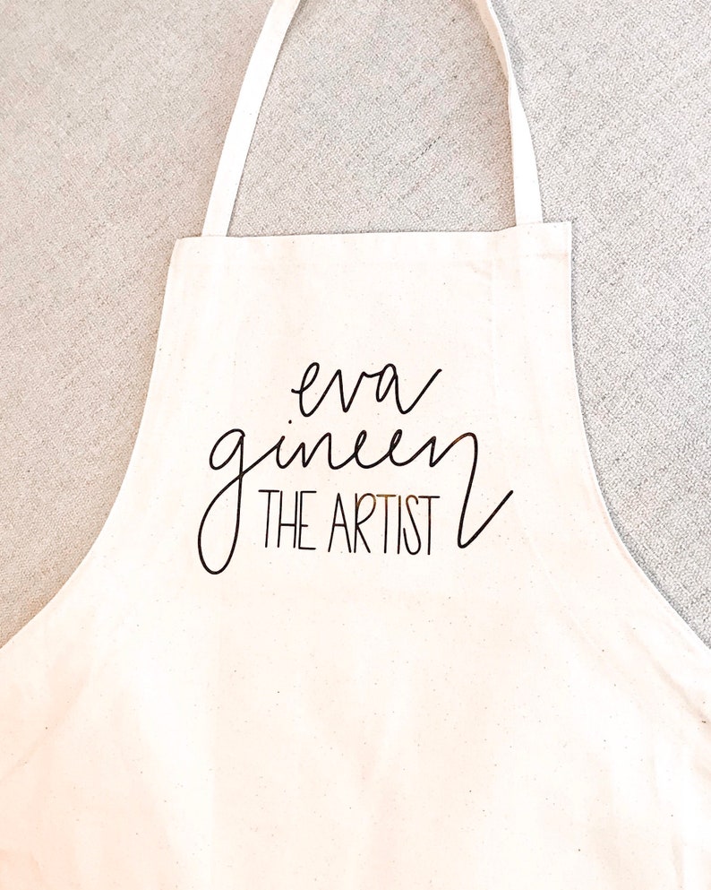 May include: White canvas apron with black text that reads "eva gineen THE ARTIST".