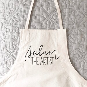 May include: White canvas apron with black text that reads "Salam The Artist".