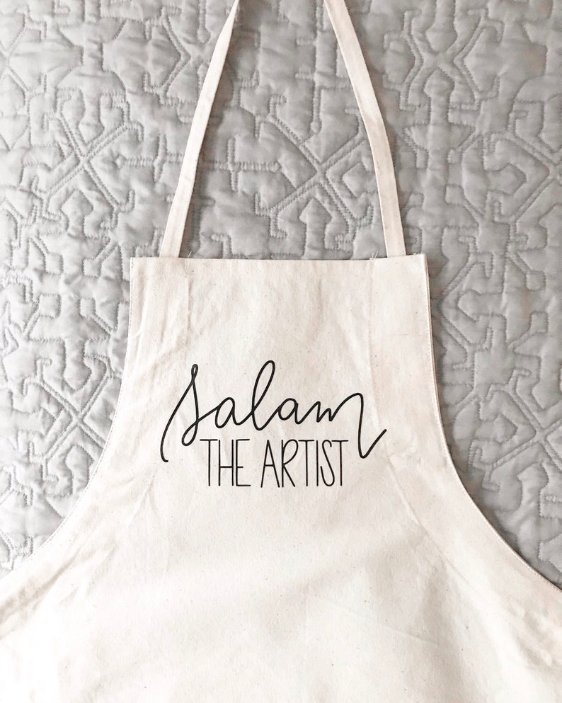 May include: White canvas apron with black text that reads "Salam The Artist".