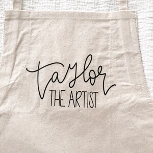 May include: A beige canvas apron with black adjustable straps. The apron has the text "Taylor The Artist" printed on the front.