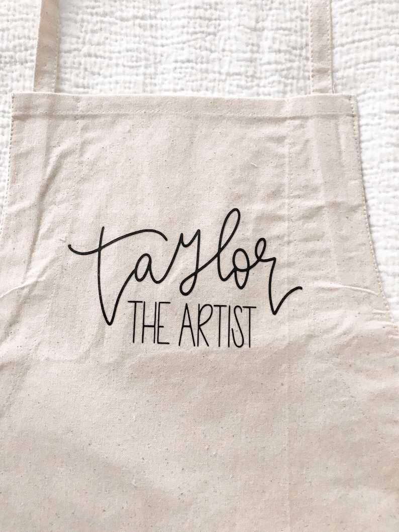May include: A beige canvas apron with black adjustable straps. The apron has the text "Taylor The Artist" printed on the front.